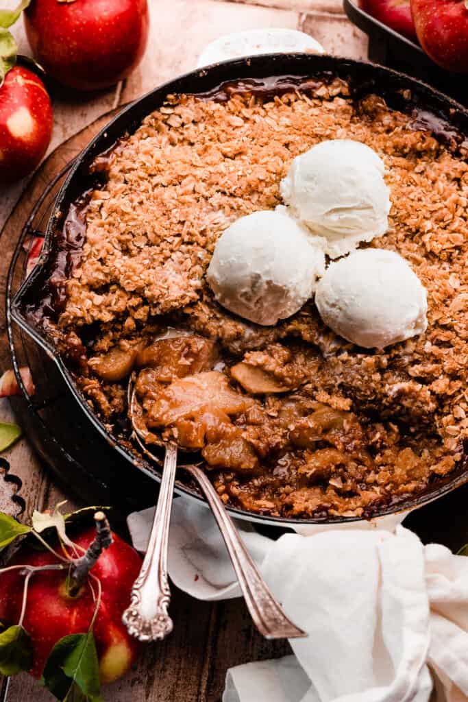 A skillet of apple crisp with scoops of ice cream and scoops of crisp missing.