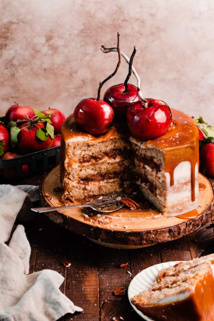 The sliced open caramel apple cake on a wooden cake stand topped with caramel drip and candy apples.