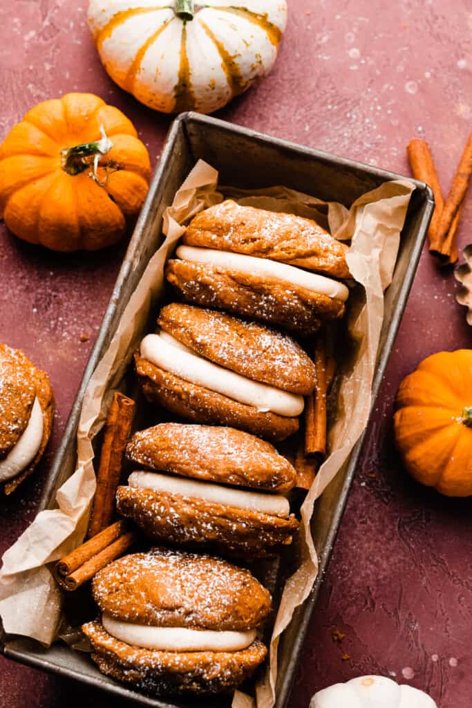 Four pumpkin whoopie pies in a loaf pan, on a burgandy backdrop with mini pumpkins scattered around.
