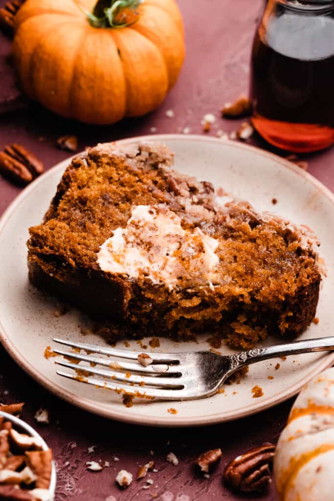 A slice of pumpkin bread on a plate, with a slather of butter and a bite missing.