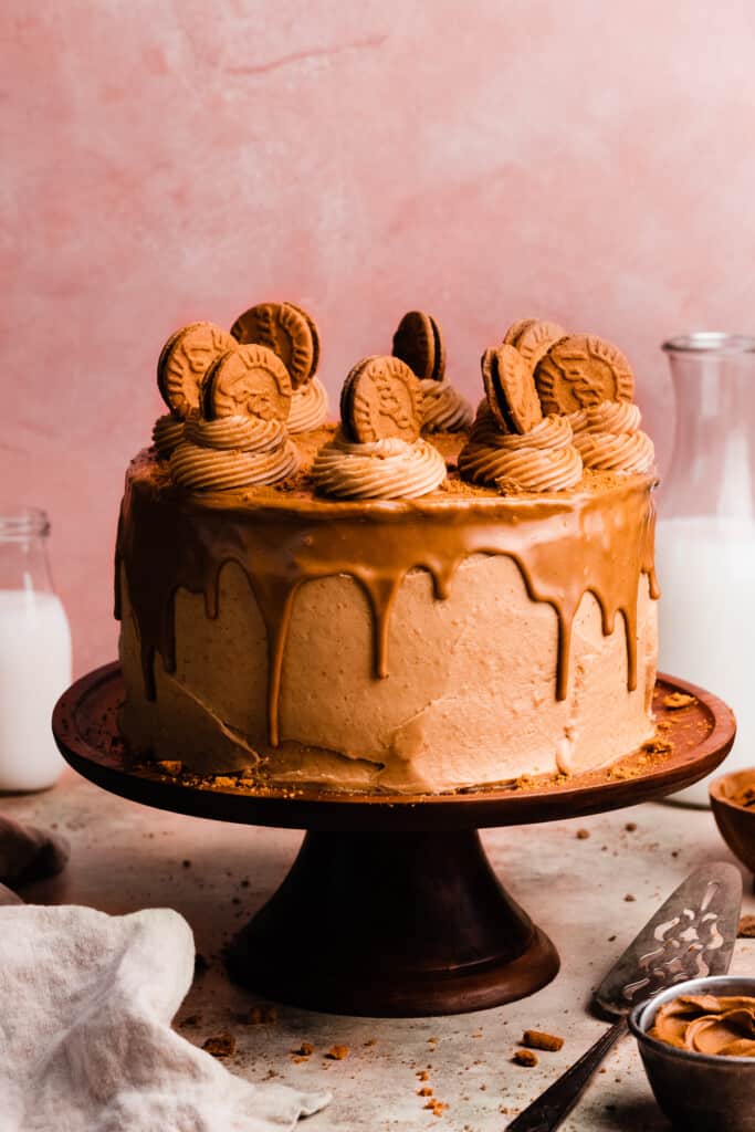 The full biscoff cake on a cake stand, topped with a cookie butter drip and frosted with swirls of cookie butter frosting topped with biscoff cookies.