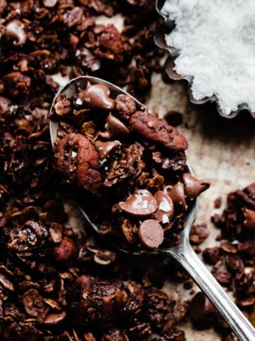 A spoonful of chocolate granola on a sheet pan, with a small dish of flaky sea salt nearby.