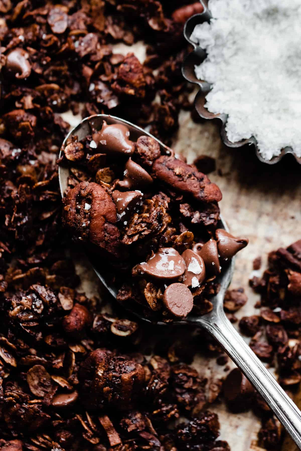 A spoonful of chocolate granola on a sheet pan, with a small dish of flaky sea salt nearby.