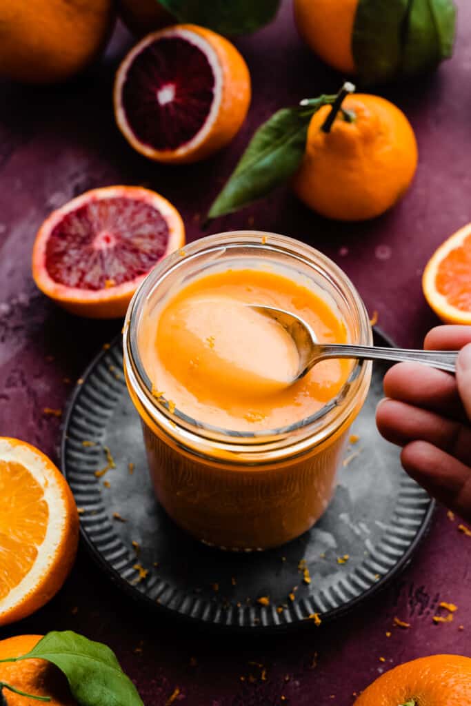 A hand dipping a spoon into the jar of orange curd.