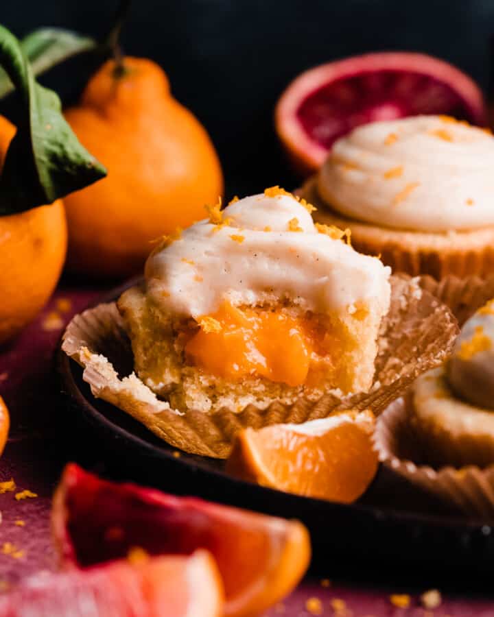 An orange cupcake on a plate, with a bite missing to reveal the orange curd filling.