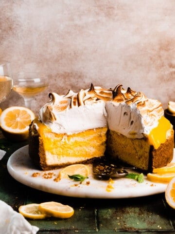 The sliced open lemon meringue cheesecake on a cake stand, showing the layers of crust, lemon cheesecake, lemon curd, and meringue topping.