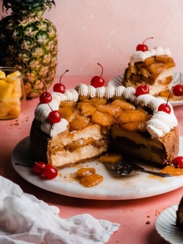 The sliced open pineapple cheesecake on a cake platter, topped with caramelized pineapple, whipped cream, and cherries.