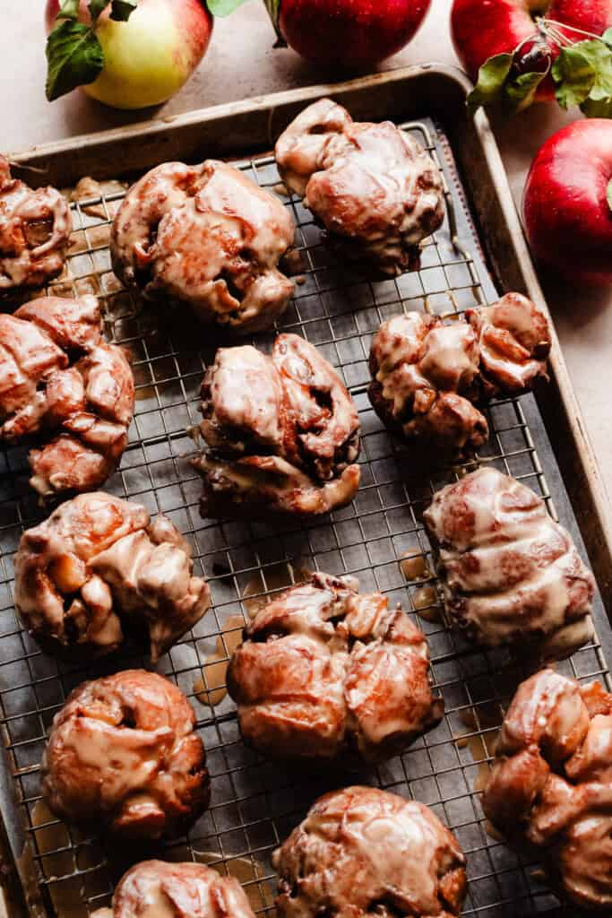 Glazed apple fritters on a cooling rack over a cookie sheet.