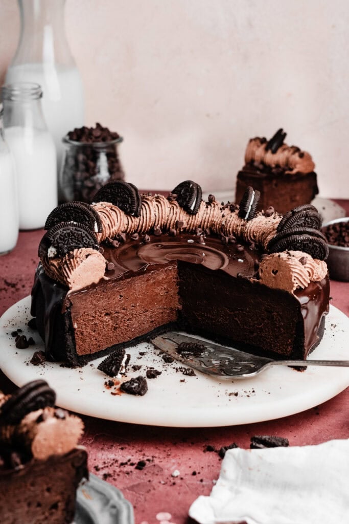 The sliced chocolate cheesecake on a cake plate, topped with chocolate ganache, chocolate whipped cream, and oreo pieces.