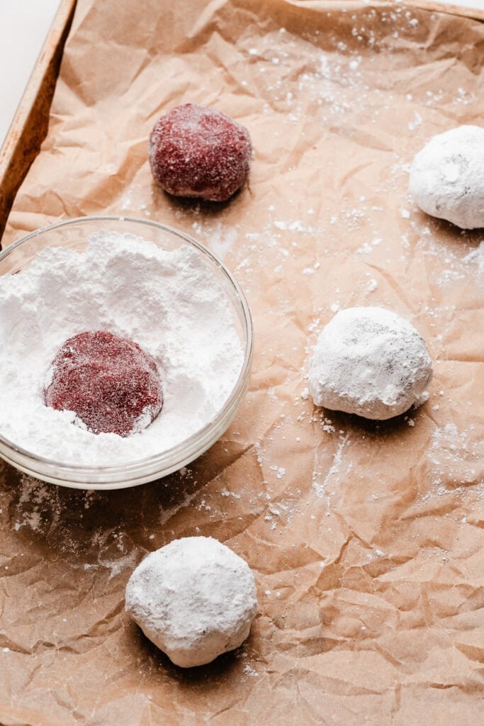 Red velvet cookie dough balls being rolled in granulated sugar and powdered sugar.