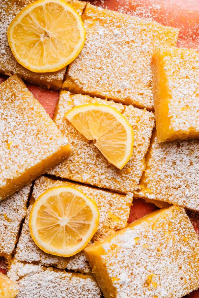 A close-up on the sliced lemon bars on a pink surface. They're dusted with powdered sugar and a few slices have lemon slices on top.