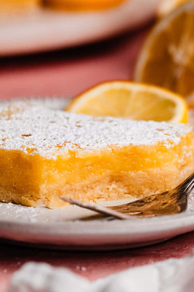 A lemon bar on a plate, dusted with powdered sugar, with a bite missing.
